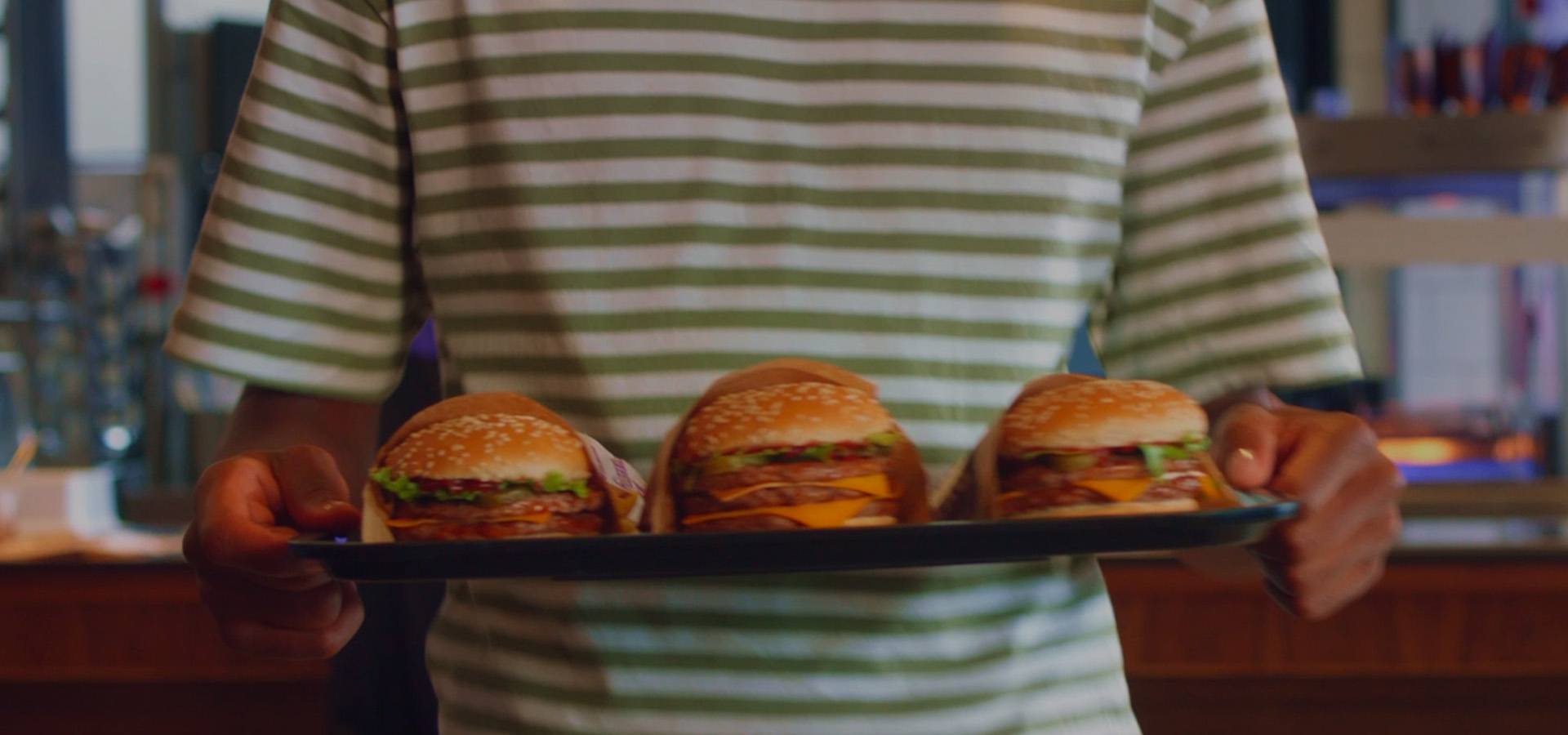 Person holding a tray of three gourmet cheeseburgers with sesame seed buns, melted cheese, lettuce and beef patties